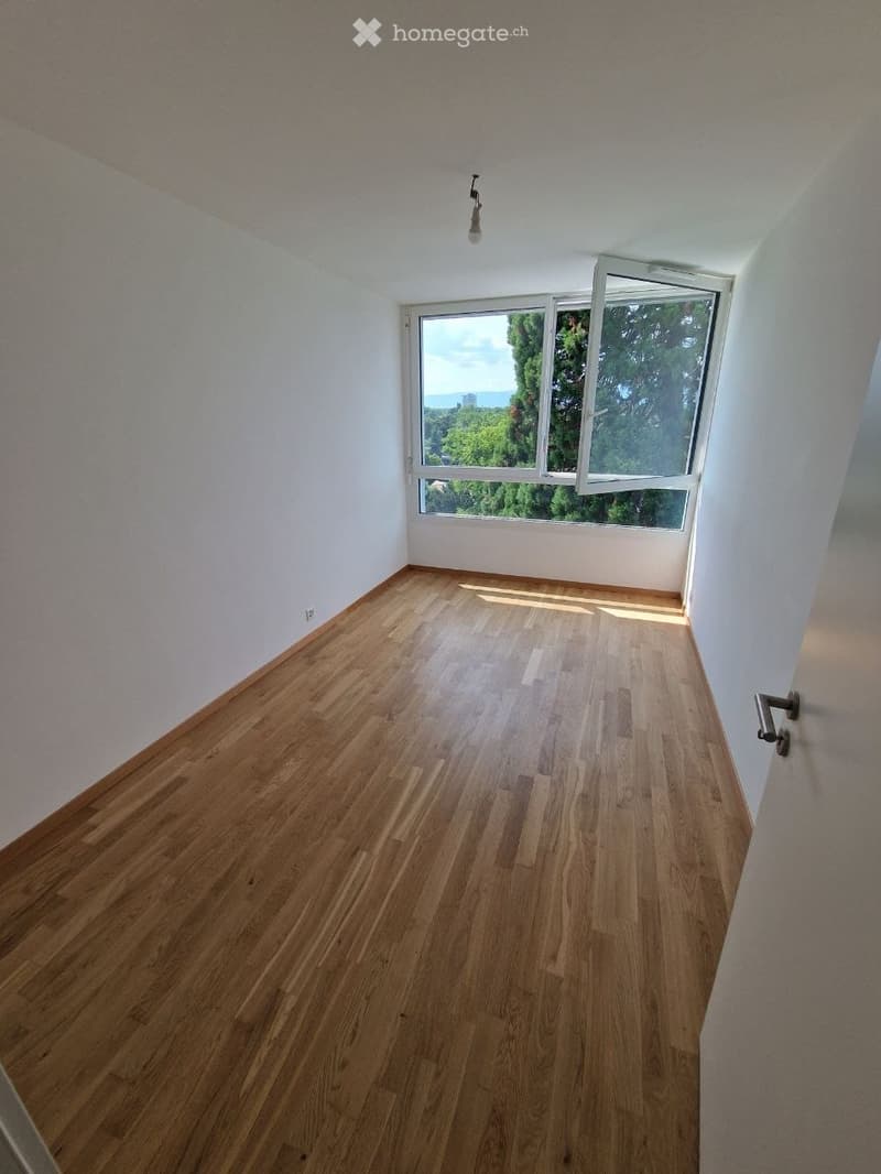 Empty room with wooden flooring, white walls, and large windows offering a view of greenery and the sea.