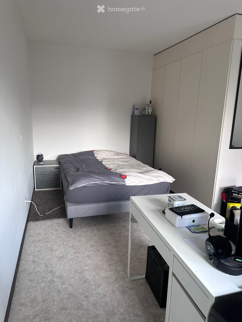 A modern, compact bedroom with a grey bed, nightstand, desk, and wardrobe. The room features a white desk with a laptop, headphones, and other small items, along with a radiator and a small nightstand with a clock.