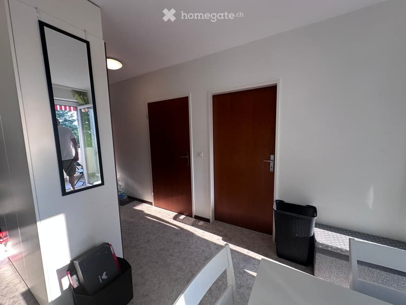 A small, well-lit hallway with white walls, a large mirror, a white chair, a laptop on a black stand, and a black trash bin.