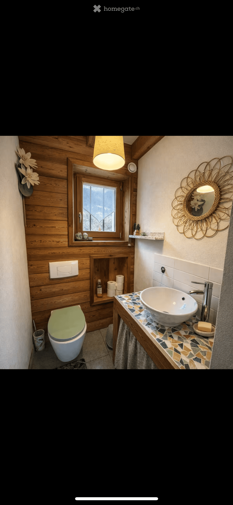 Modern bathroom with wooden walls, a sink with a decorative faucet, a toilet, a large mirror with a sun-shaped frame, and a window with a view.