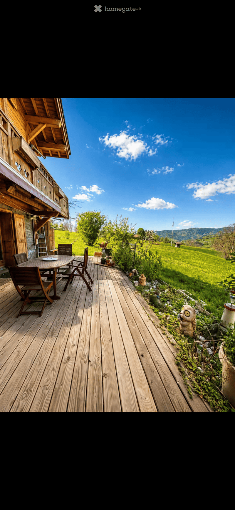 Wooden deck with a table and chairs, overlooking a green field and mountains under a clear blue sky.