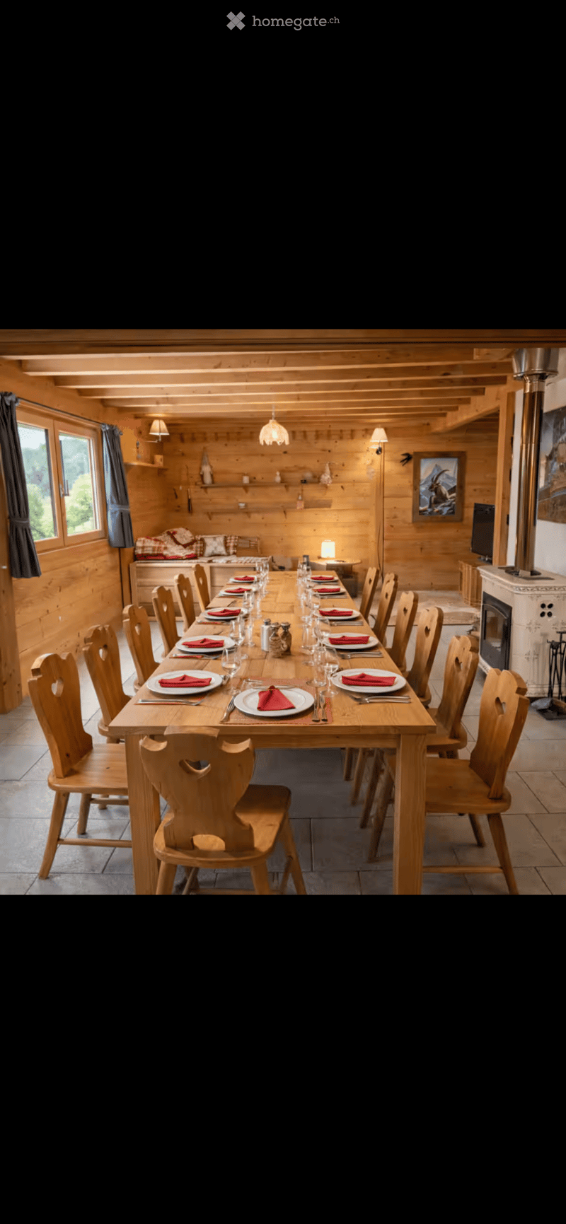 A cozy, wooden-paneled dining room with a long wooden dining table set for multiple guests, surrounded by wooden chairs. A fireplace is visible on the right, and a small seating area with a sofa and lamp is in the background.