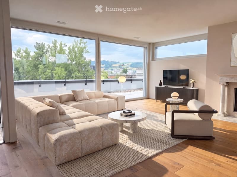 Modern living room with large windows offering a scenic view, a beige sectional sofa, a coffee table, an armchair, a television, a floor lamp, and a fireplace.