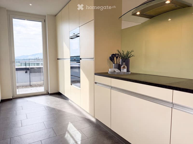 Modern kitchen with light-colored cabinets, a built-in oven, and a countertop with kitchen utensils and a decorative plant. Large window offers a view of the outdoor area.