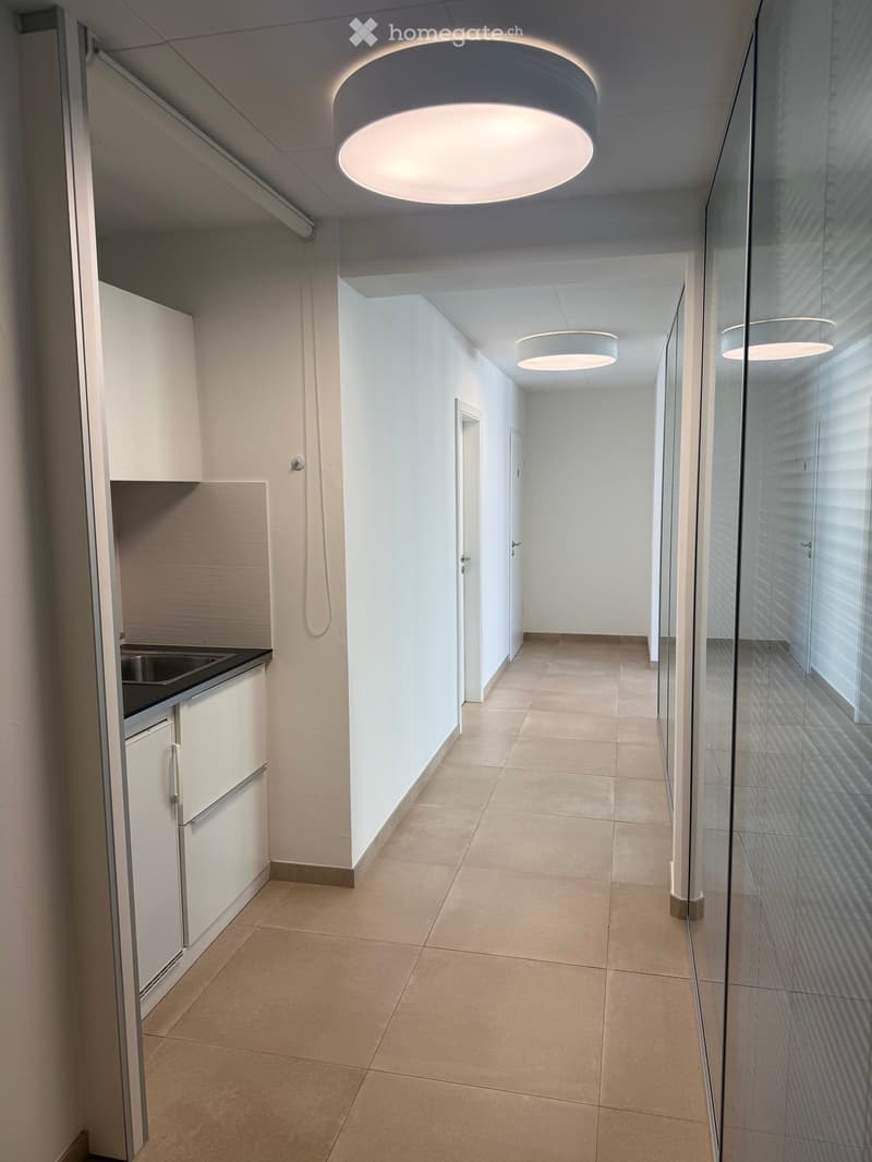 Modern hallway with white walls, beige tiled floor, and recessed lighting. Features a glass wall on one side and a small kitchenette area on the left.