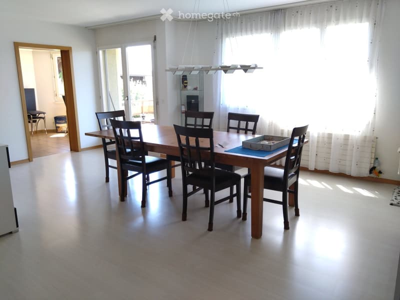 Bright dining room with a wooden dining table surrounded by six black chairs, large windows with white curtains, and a hanging light fixture above the table.