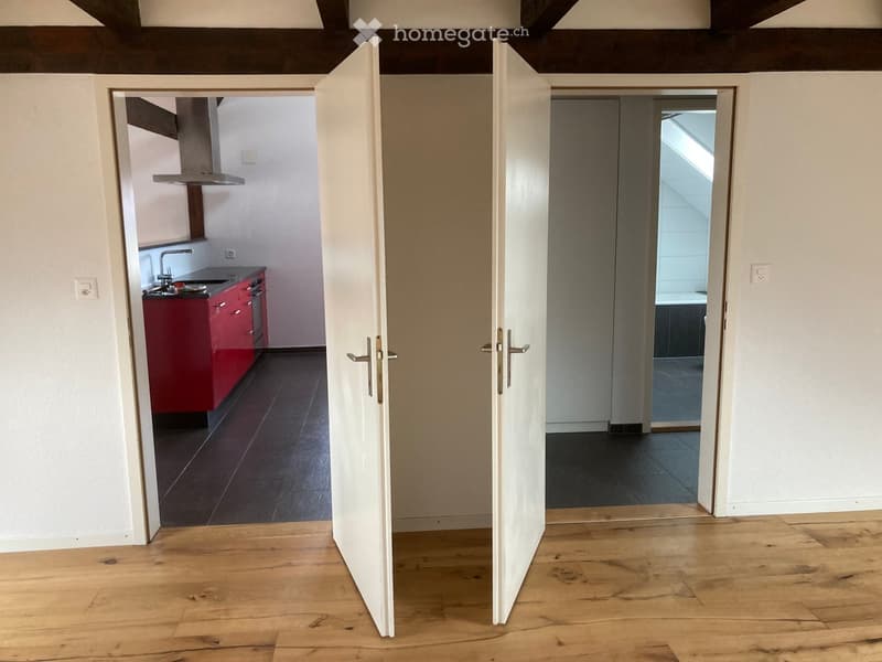Open hallway with wooden flooring, leading to a kitchen and a bathroom. The kitchen features red cabinets and a sink, while the bathroom has a visible bathtub and tiled walls.