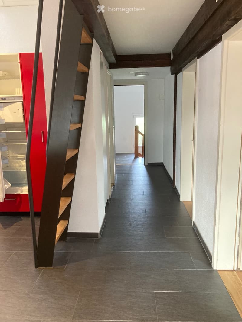 Modern hallway with dark tiled flooring, white walls, and wooden staircase leading upwards. Features include built-in wooden shelving on one side and a red accent wall in the adjacent room.