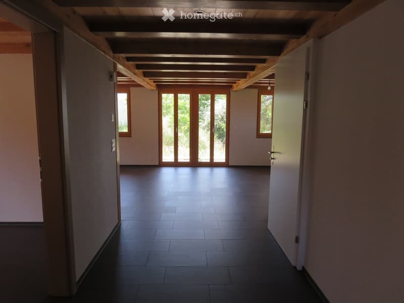 Empty hallway with wooden ceiling beams, dark tiled floor, and large glass doors leading outside.