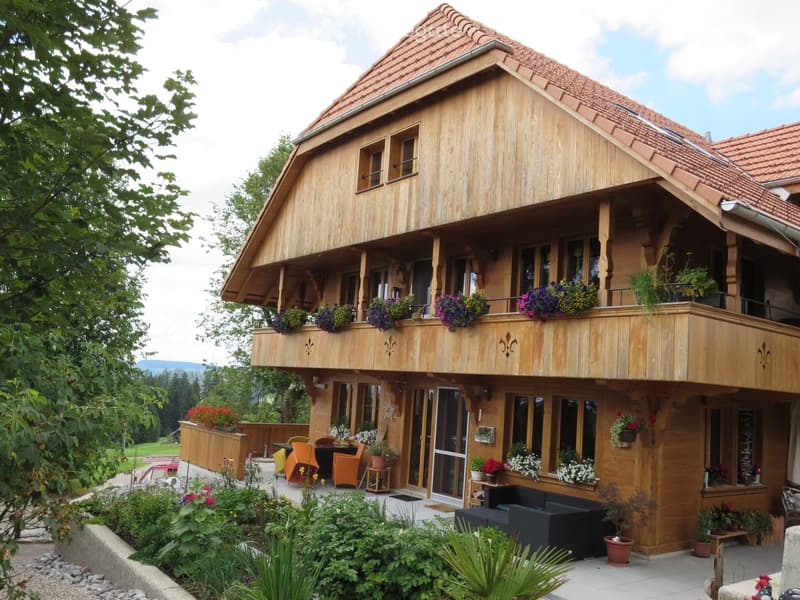 Two-story wooden house with a red-tiled roof, large balcony, and well-maintained garden with various plants and seating.