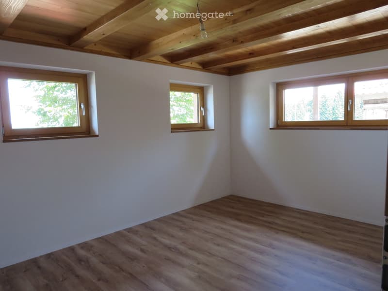 Empty room with light wooden flooring, white walls, and three windows with wooden frames. The ceiling features exposed wooden beams.