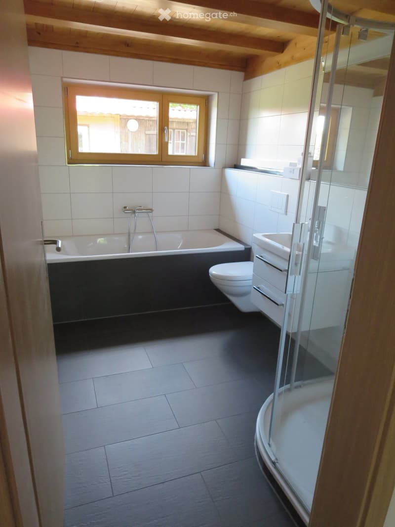 Modern bathroom with a bathtub, toilet, sink, and a glass-enclosed shower. The room features white tiles, a dark floor, and a wooden-framed window letting in natural light.