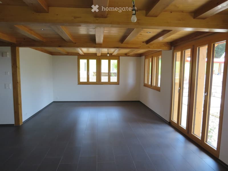 Empty room with dark tiled floor, light-colored walls, wooden ceiling beams, and large windows with wooden frames. No furniture or equipment present.