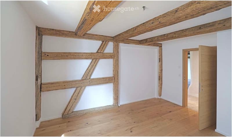 Empty room with exposed wooden beams and light wooden flooring, featuring white walls and a doorway leading to another space.