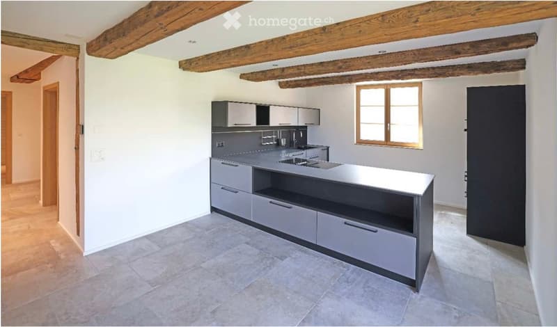 Modern kitchen with grey cabinets, a central island, and a stainless steel sink. Exposed wooden beams on the ceiling and a large window provide natural light.