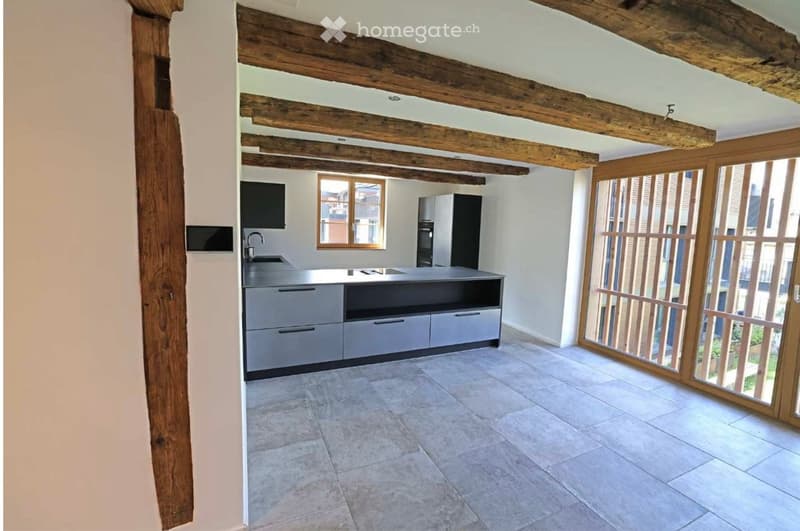 Modern kitchen with exposed wooden beams, grey cabinets, a central island, and large windows with wooden shutters.
