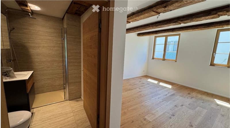 Empty room with wooden flooring, white walls, and exposed wooden beams on the ceiling. A separate bathroom with a sink, toilet, and glass shower enclosure is visible through an open doorway.