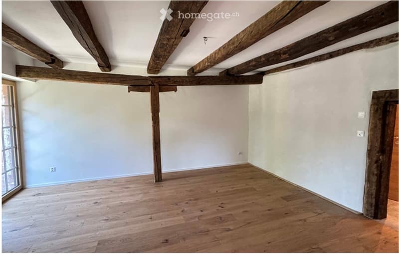 Empty room with exposed wooden beams on the ceiling, wooden flooring, and white walls. No furniture or equipment present.