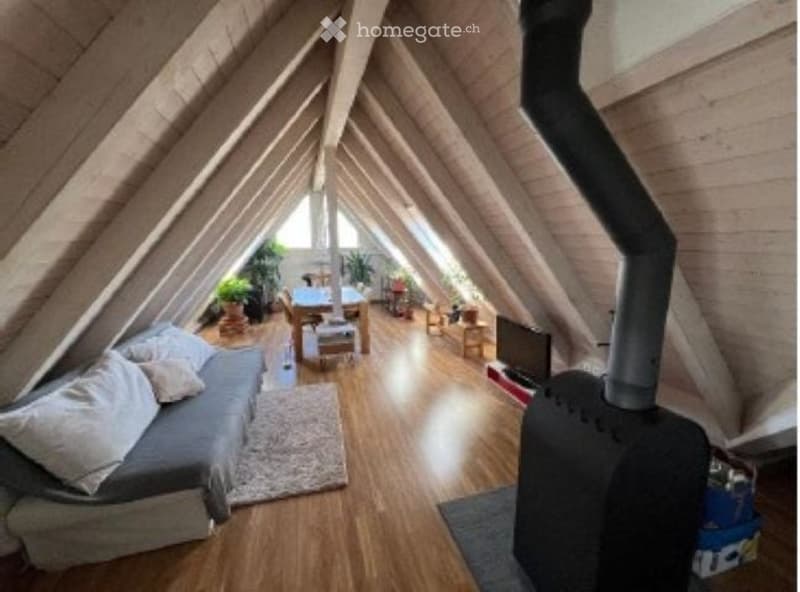 Cozy attic space with wooden beams, exposed wooden floor, grey sofa, coffee table, dining area with table and chairs, television, and a fireplace.