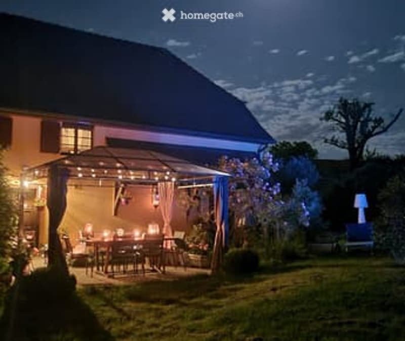 Nighttime view of a well-lit outdoor patio area with string lights, a dining table, and chairs, surrounded by greenery and a house in the background.