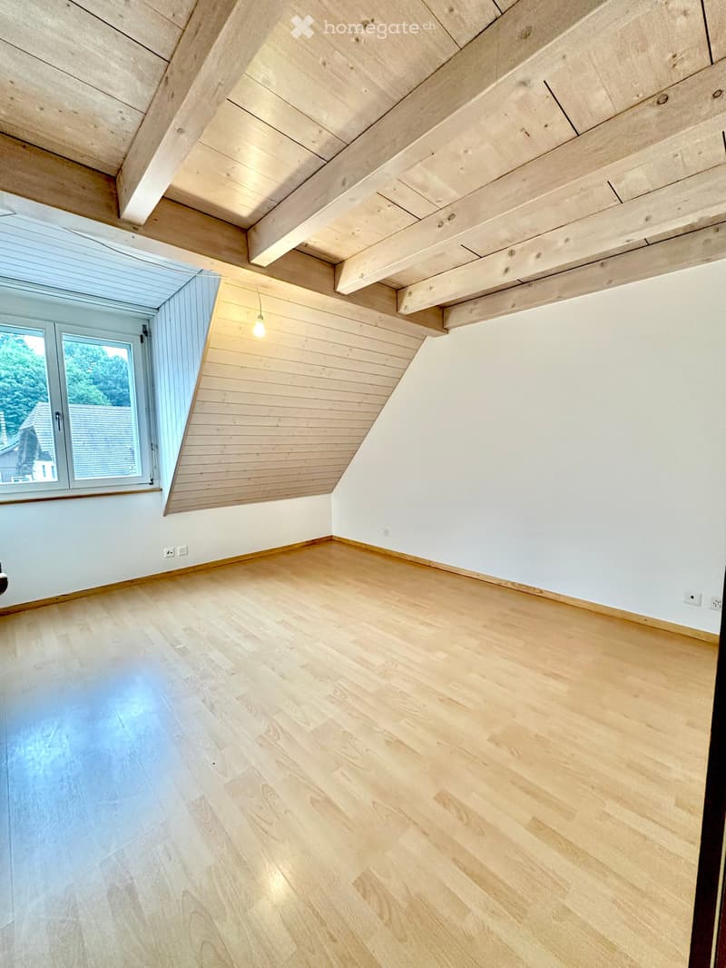 Empty room with wooden flooring and exposed wooden beams on the ceiling, featuring a large window with a view of greenery.