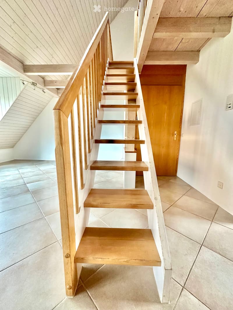Modern staircase with wooden steps and handrail, leading to an upper level. The walls are white, and the floor is tiled.