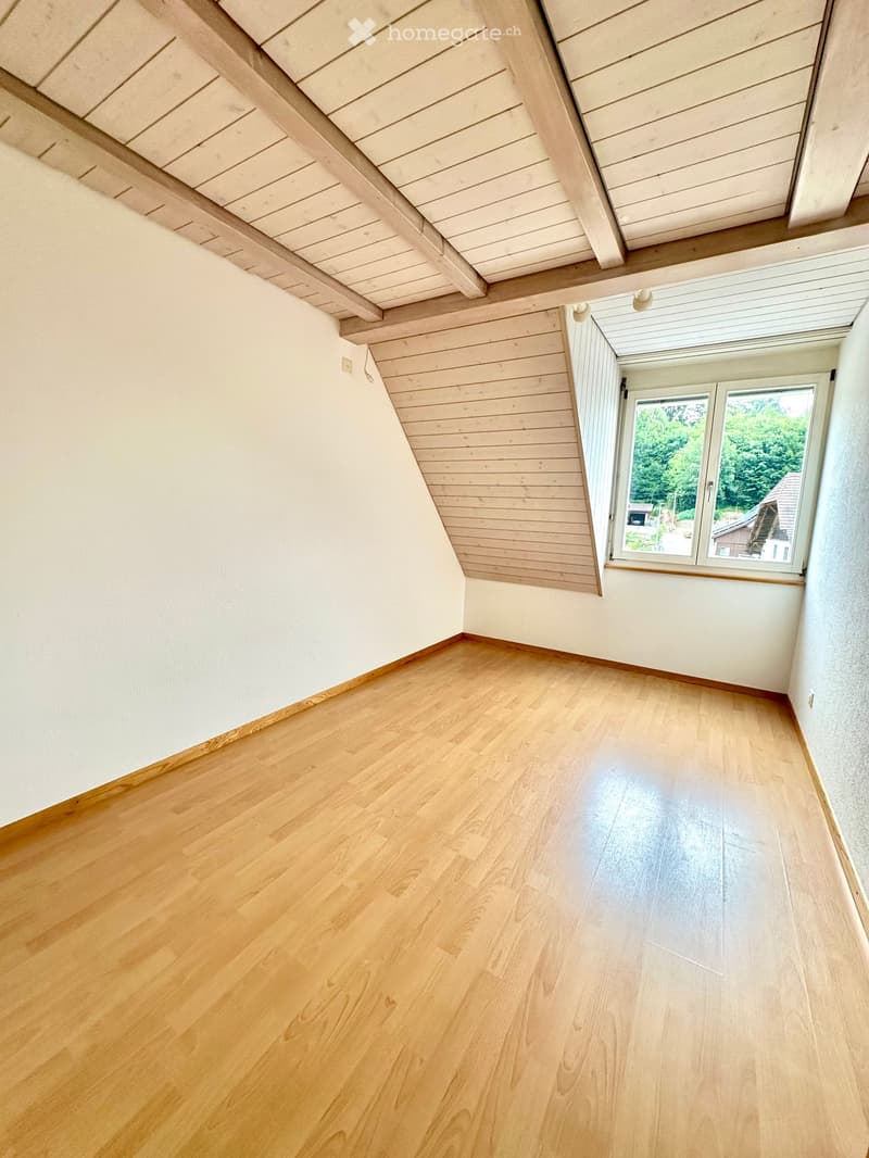 Empty room with wooden flooring, white walls, and a slanted wooden ceiling. A large window with white frames offers a view of greenery outside.