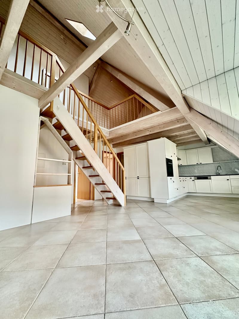 Open-plan space with a staircase leading to an upper level, modern kitchen area with white cabinets and an oven, and tiled flooring.
