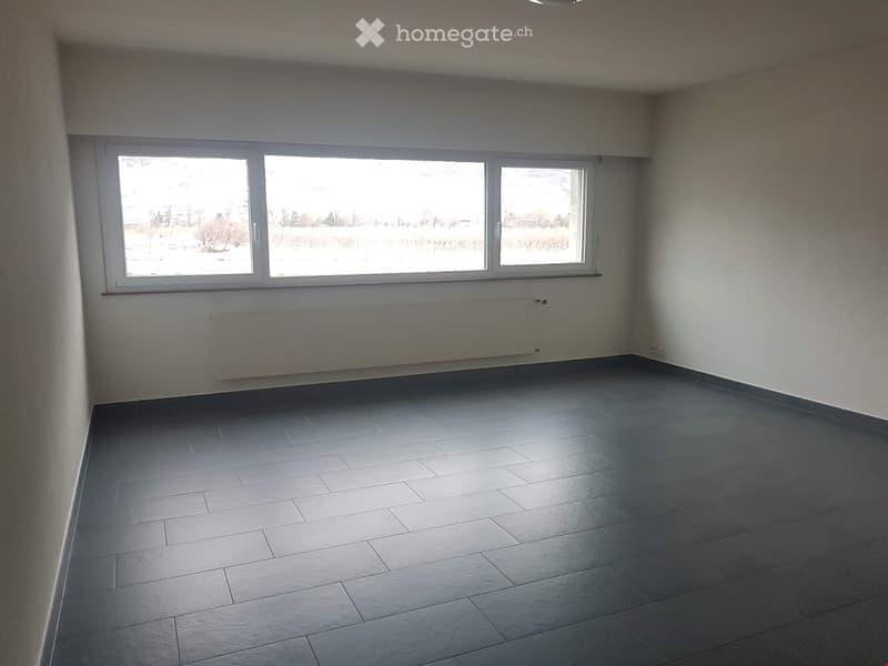 Empty room with grey tiled floor, white walls, and three large windows allowing natural light.