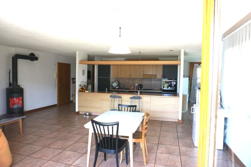 The image shows a modern, well-lit kitchen and dining area. The kitchen features light wooden cabinets, a white countertop, and various appliances including a refrigerator and an oven. A white dining table with four chairs is positioned in the foreground.