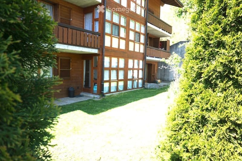 A multi-story wooden building with large windows and balconies, surrounded by greenery and a well-maintained lawn.