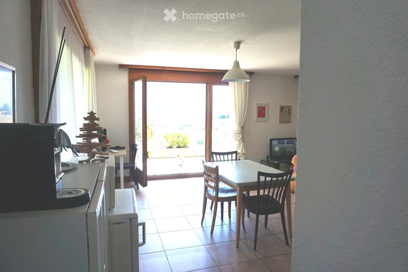 Bright living room with a dining table, chairs, refrigerator, TV, and large windows leading to a balcony.