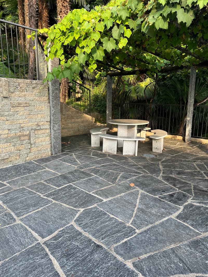 Outdoor patio with stone tiles, a round stone table, and benches surrounded by lush greenery and a metal railing.