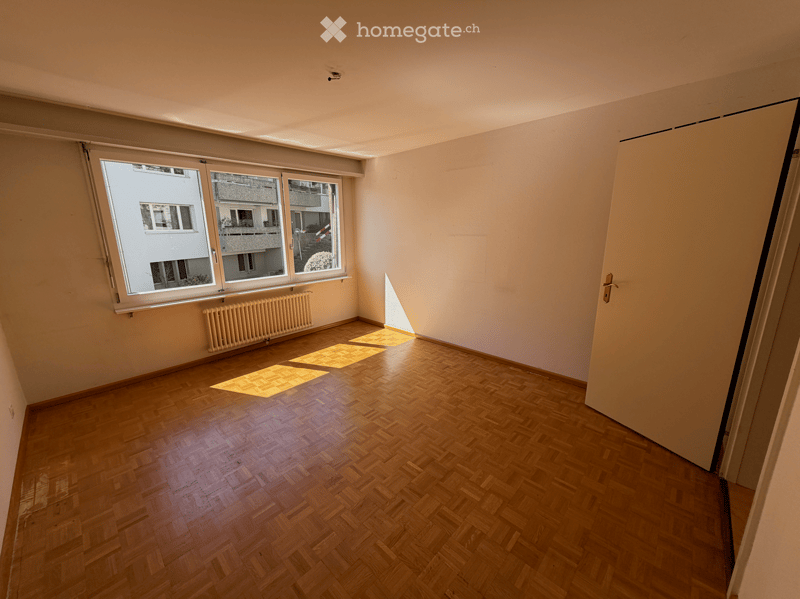 Empty room with wooden flooring, large windows, radiator, and a door leading to another space.