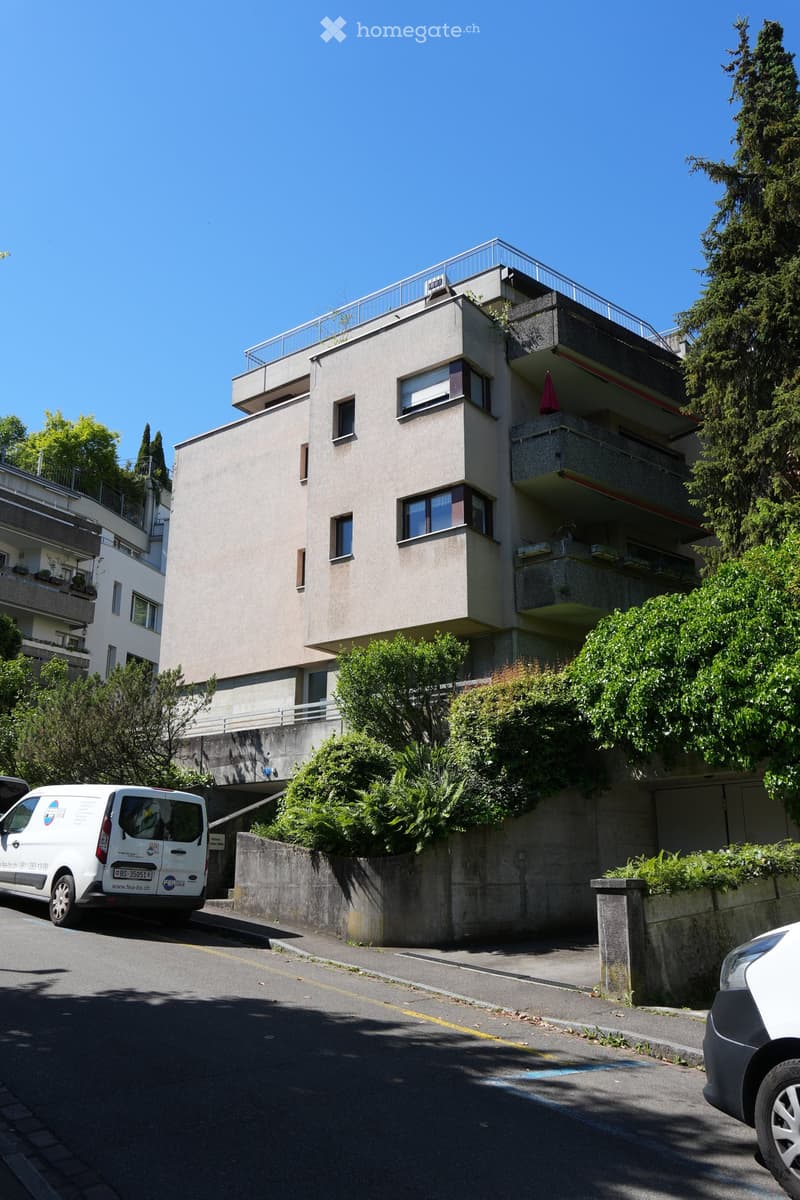Modern multi-story apartment building with balconies, surrounded by greenery and trees. A white van is parked on the street in front of the building.
