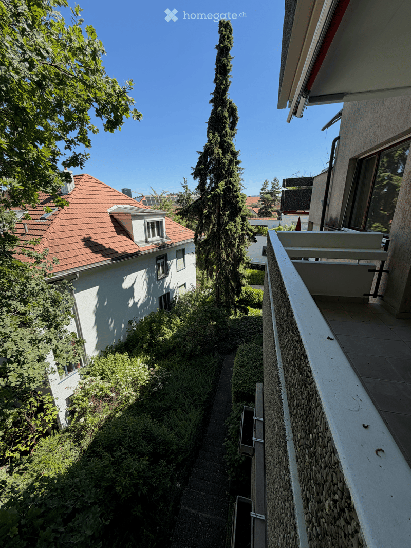 Balcony with stone balustrade, overlooking a narrow alleyway lined with greenery and neighboring houses.