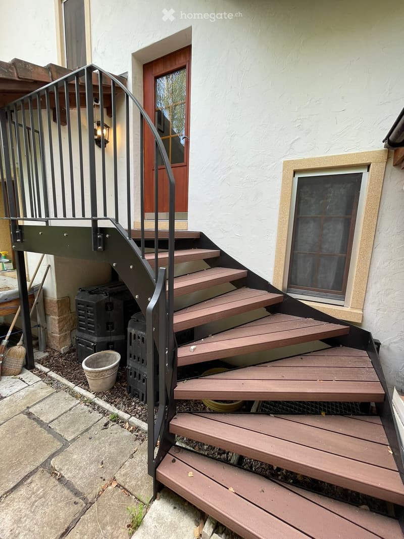 Wooden staircase leading up to a house entrance with a metal railing, a small planter, and a broom leaning against the wall.