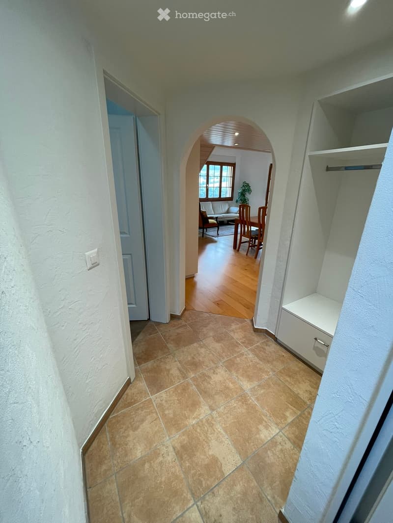 A well-lit hallway with tiled flooring, white walls, and an arched opening leading to a dining area with wooden furniture and a plant.