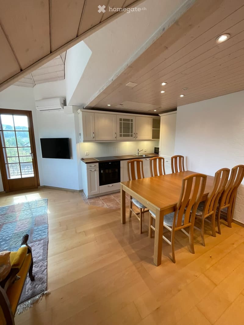 Open-plan kitchen and dining area with wooden flooring, white cabinets, a dining table with six chairs, and a screen mounted on the wall.