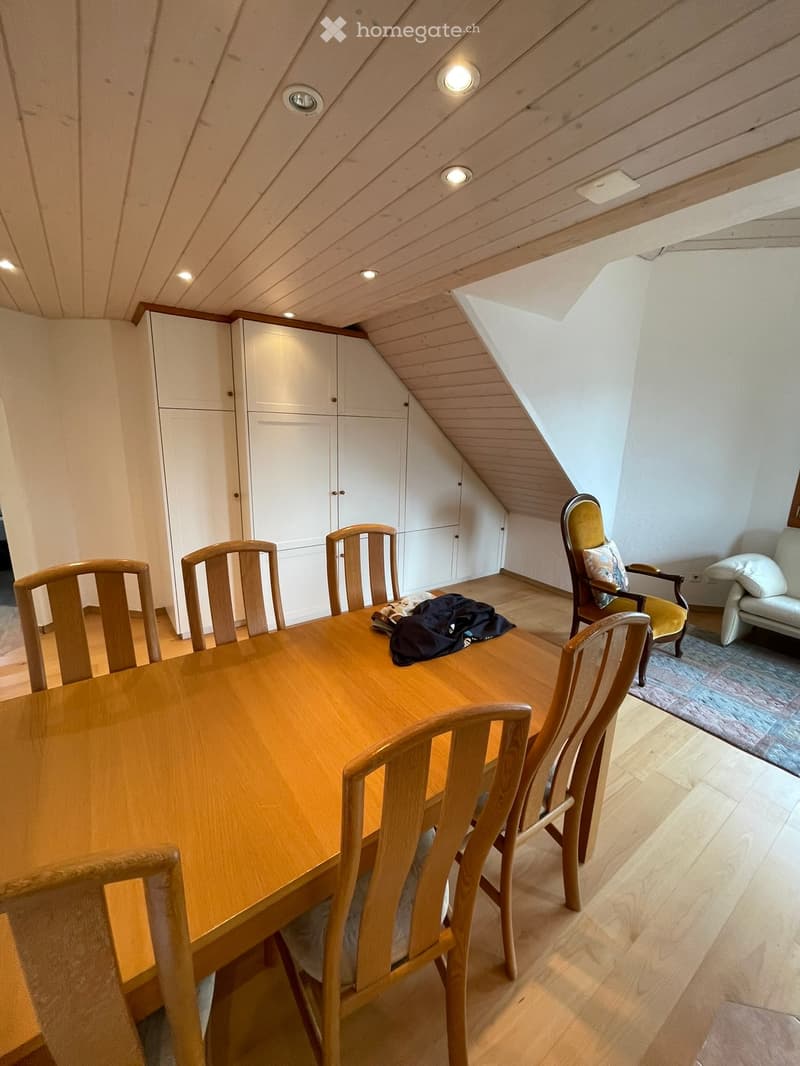 Bright attic room with wooden table, chairs, sofa, and built-in white cabinets under a slanted wooden ceiling with recessed lighting.