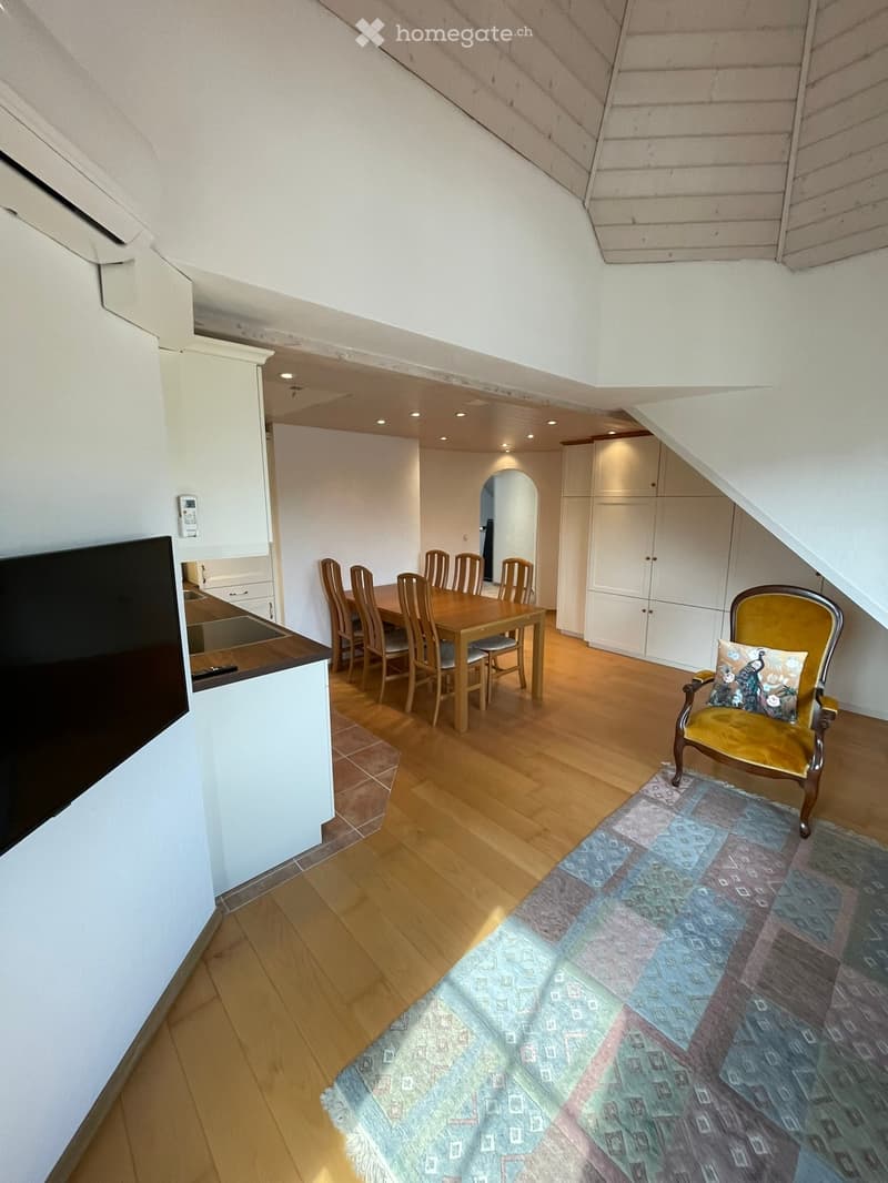A cozy attic room with wooden flooring, a dining area with a table and chairs, a television, and a decorative armchair with a patterned cushion on a colorful rug.