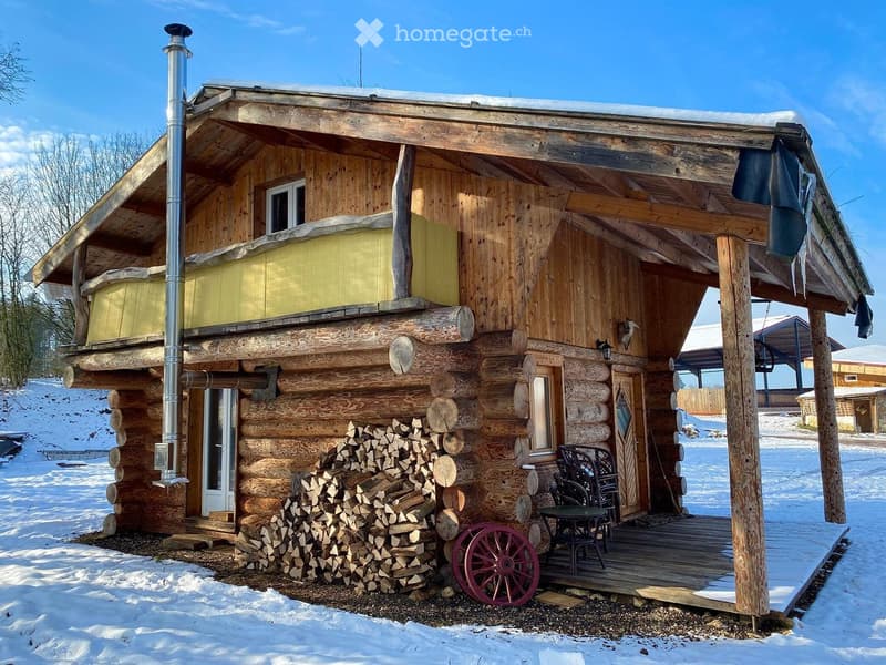 A rustic wooden log cabin with a raised ground floor, chimney, and a porch with outdoor furniture.