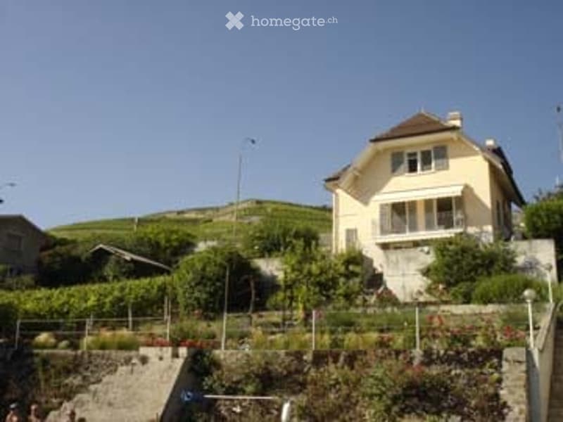 A two-story yellow house with white trim, situated on a hillside with a vineyard in the background. The house features a balcony and is surrounded by greenery and a stone retaining wall.