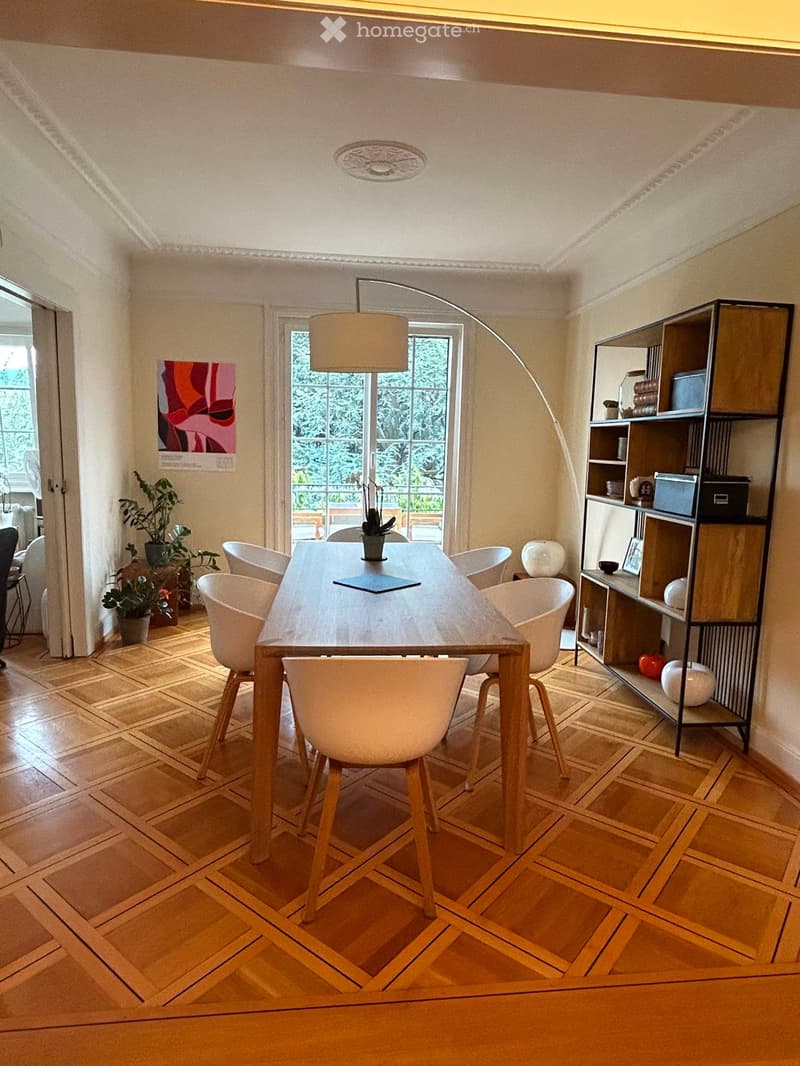 Dining room with wooden herringbone floor, long wooden dining table surrounded by white chairs, modern shelving unit with decorative items, and large windows with view of greenery.