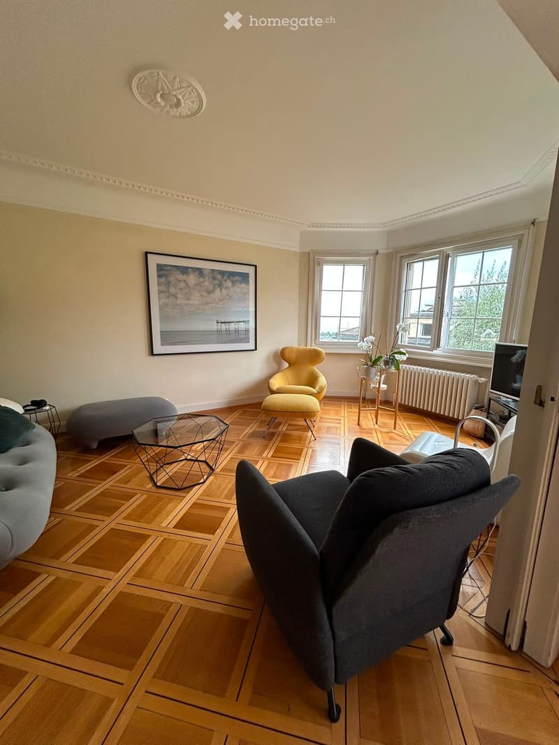 Living room with wooden flooring, large windows, a grey sofa, a yellow armchair, a glass coffee table, a small table with a plant, and a television on a stand.