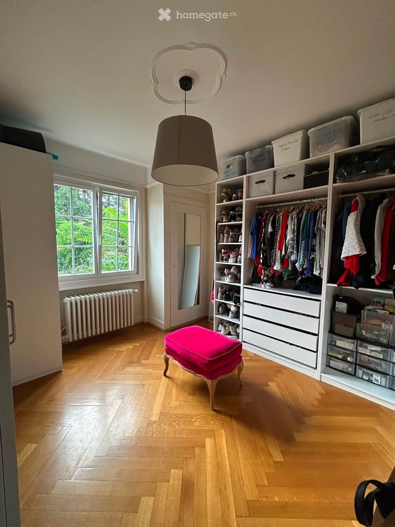 Well-lit room with wooden flooring, large window, built-in wardrobe with hanging clothes and shelves, storage boxes, a pink stool, and a mirror.