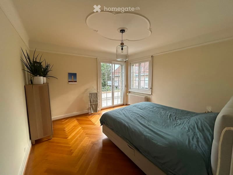 Bedroom with wooden floor, large window, bed with blue duvet, wardrobe, plant, picture frame, radiator, and a hanger.
