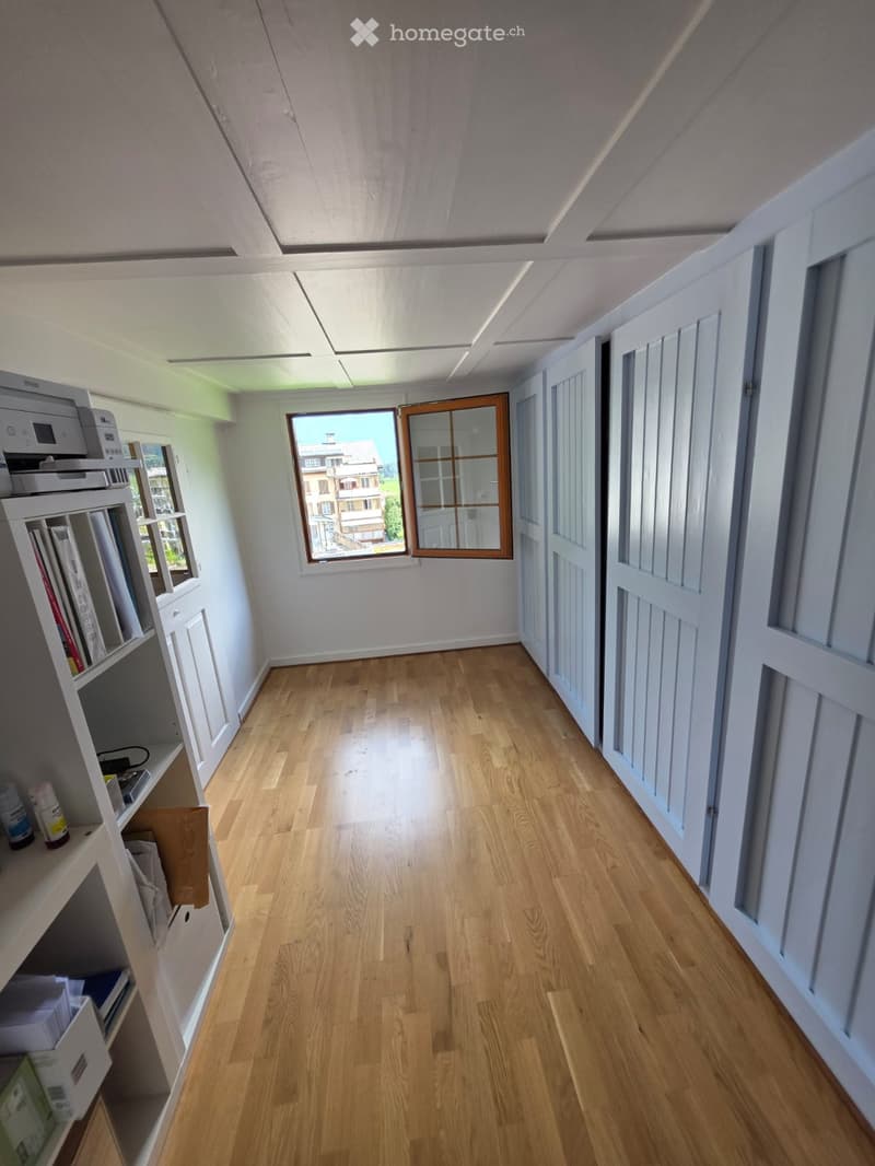 Empty hallway with wooden flooring, white walls, and several doors. A window at the end provides natural light, and a shelf with books and a printer is visible on the left side.