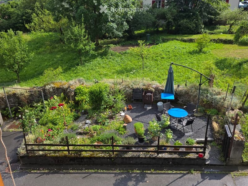 A small, well-maintained garden with a seating area featuring a blue table, chairs, and an umbrella. The garden includes various potted plants and is enclosed by a wooden fence.
