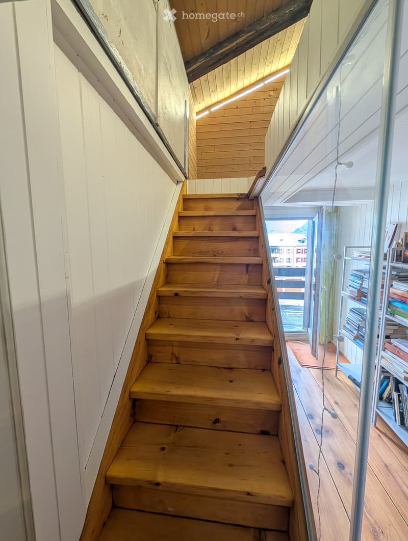 Wooden staircase with white walls and a glass railing, leading to a room with bookshelves and a view of the outside.